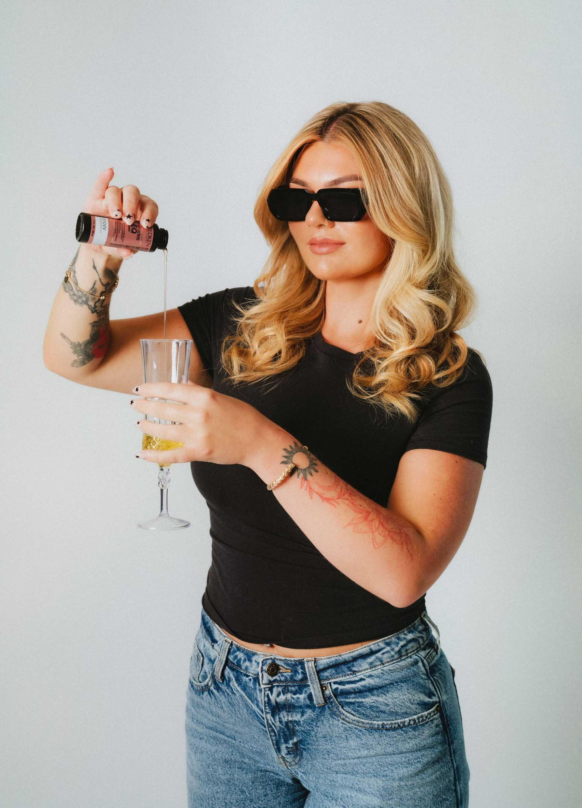 A woman pours a drink into a glass, wearing sunglasses and stylish casual attire.