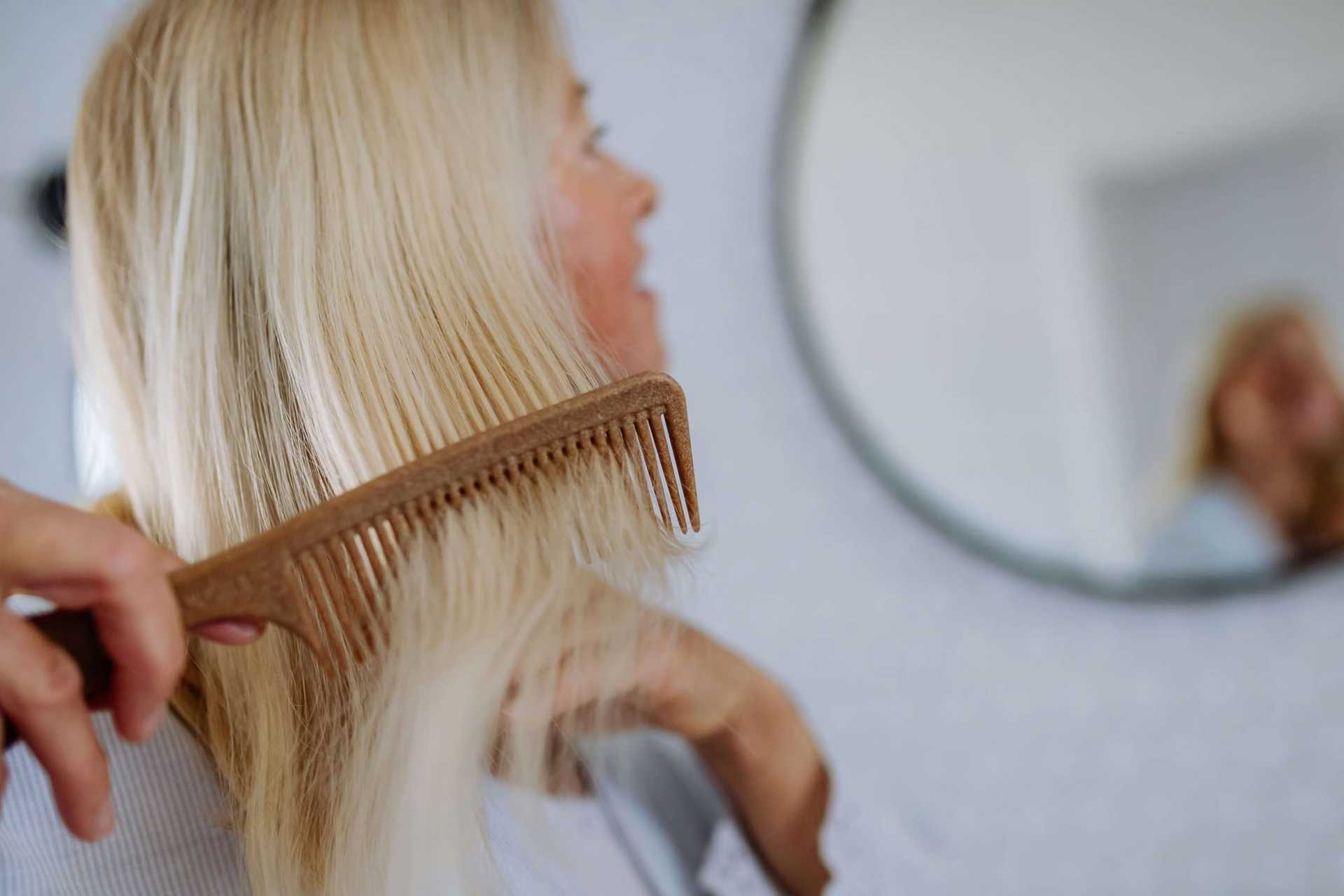 Blonde person combing hair in front of a round mirror.