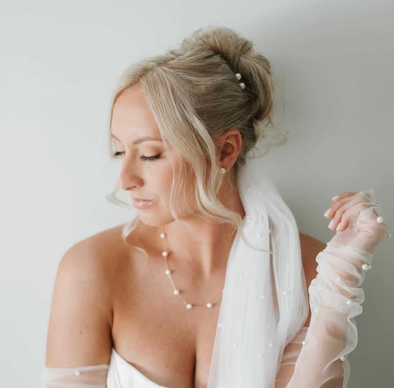 Bride in white dress and pearl accessories, gazing down, with elegant hairstyle and sheer gloves.